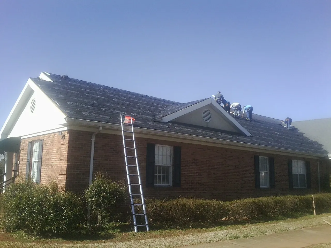 Crew tearing shingles off a brick ranch roof with a ladder set — Metal Roof Repair work by our crew in Mchenry