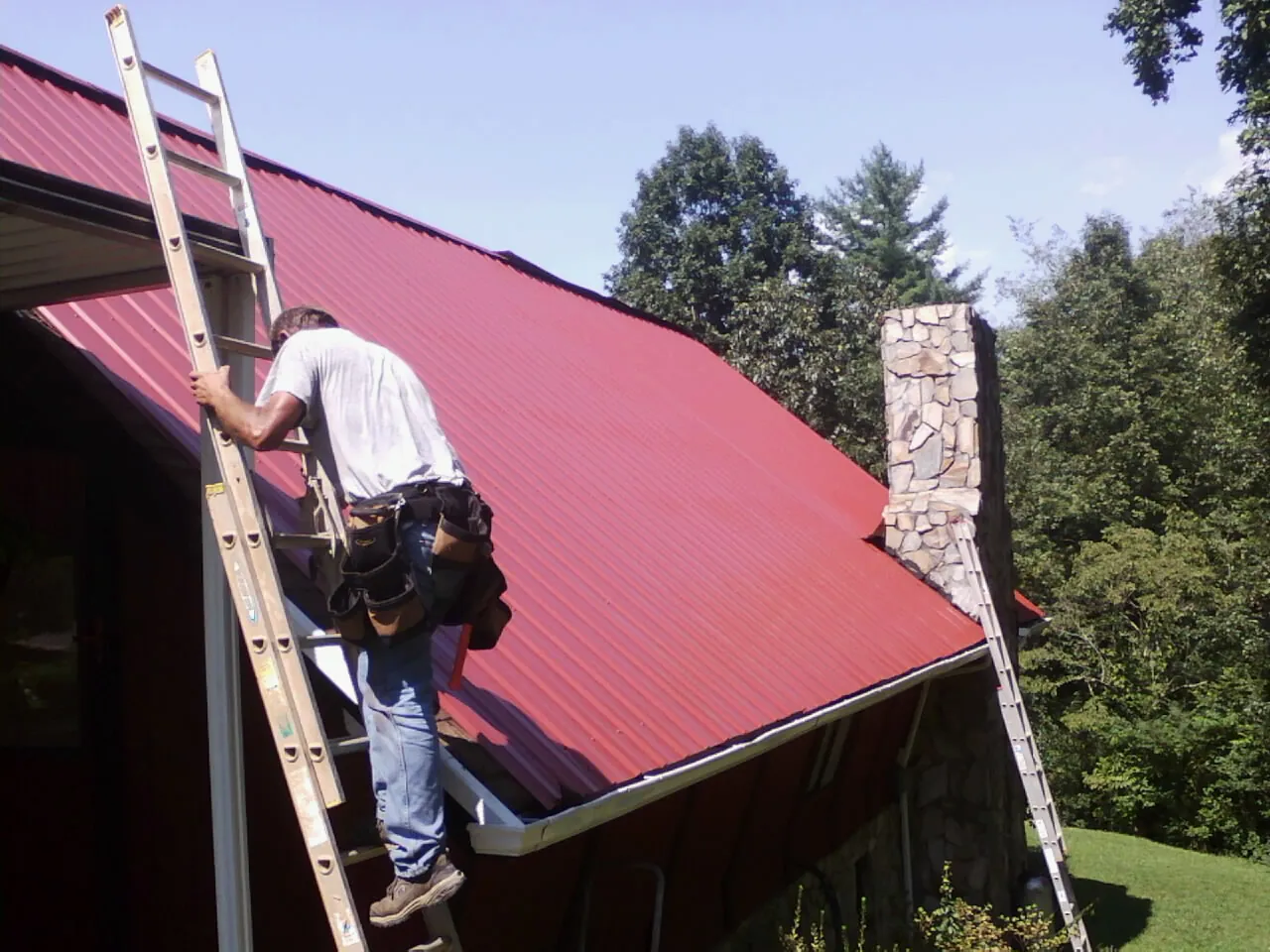 Roofer on a ladder beside a red metal roof and stone chimney — Soffit & Fascia Repair by Roofing Brothers in Mchenry